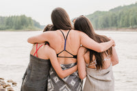 three women with their arms around each other wrapped in 100% ultra-soft Turkish towels. Each towel is being worn as a beach wrap/sarong. One towel is Aztec print grey and white and the other two are grey and beige solid colours with a nice classic weave.